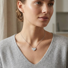 A woman wearing a delicate silver chain necklace with a small, sparkling heart-shaped pendant, styled with a light gray V-neck sweater in a softly lit indoor setting.