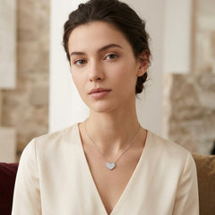 A woman wearing a delicate silver heart pendant necklace with a pavé diamond center, styled with a cream-colored satin blouse in a softly lit indoor setting.