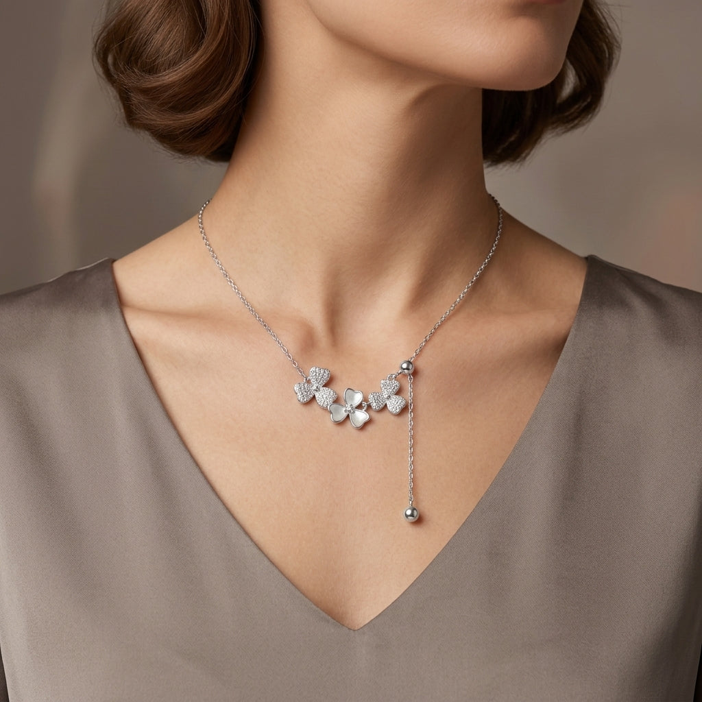 A close-up of a woman's neck and upper chest wearing a delicate silver necklace with a cluster of small flower-shaped charms and a dangling bead, paired with a taupe V-neck top.